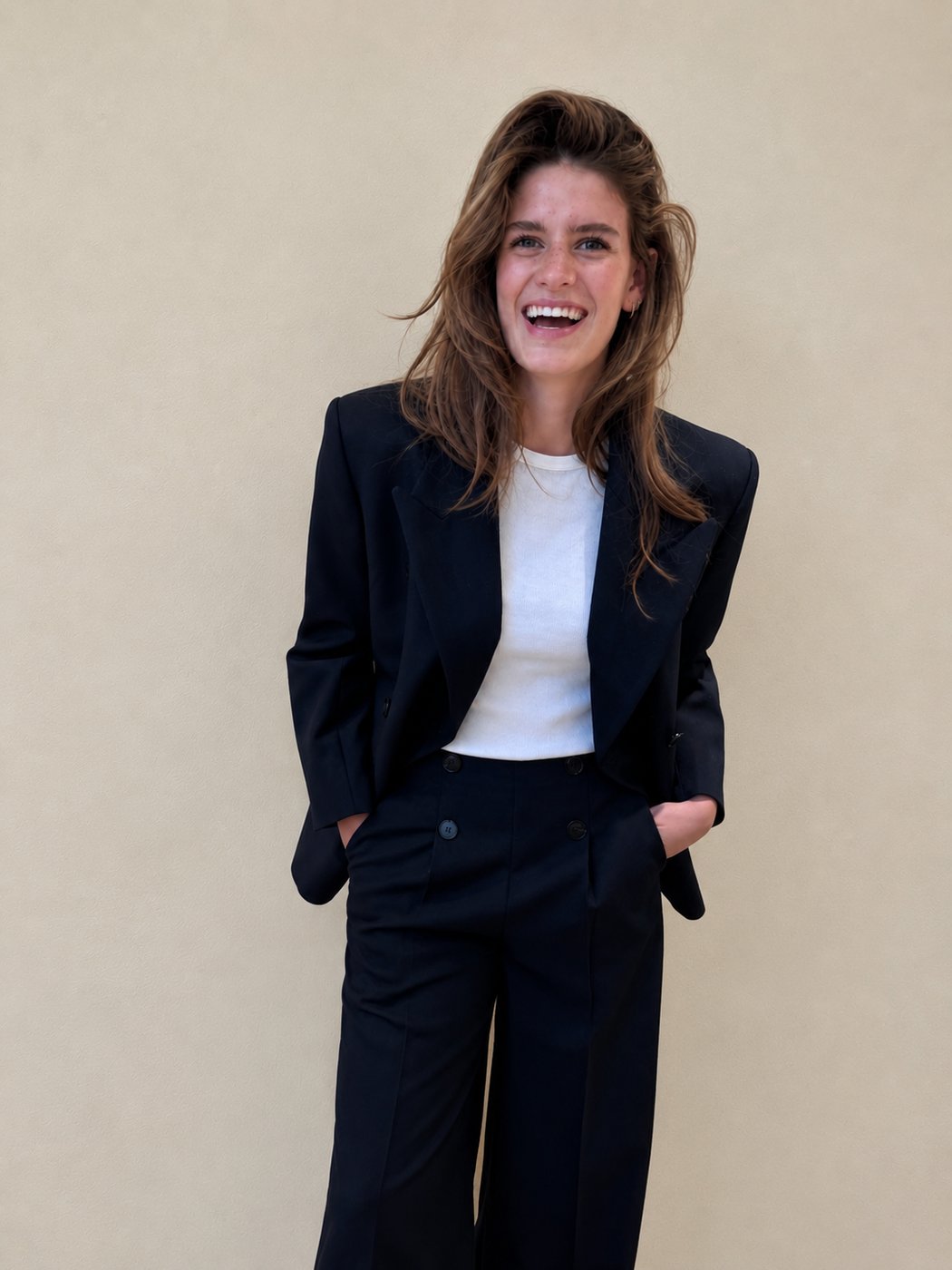 Elin laughing, wearing a tailored black blazer and trousers over a white top, against a plain warm-beige backdrop