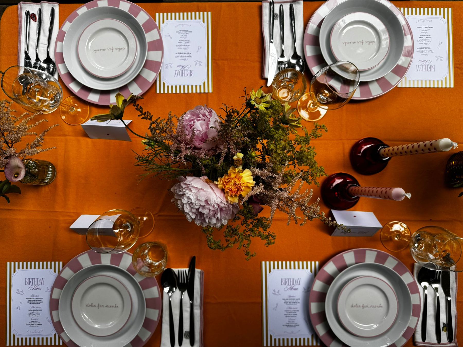 Overhead view of a symmetrical table setting on a mustard tablecloth, with pink-striped charger plates printed "dolce far niente", a loose peony and wildflower centrepiece, ruby glass candle holders and gold-striped birthday menu cards