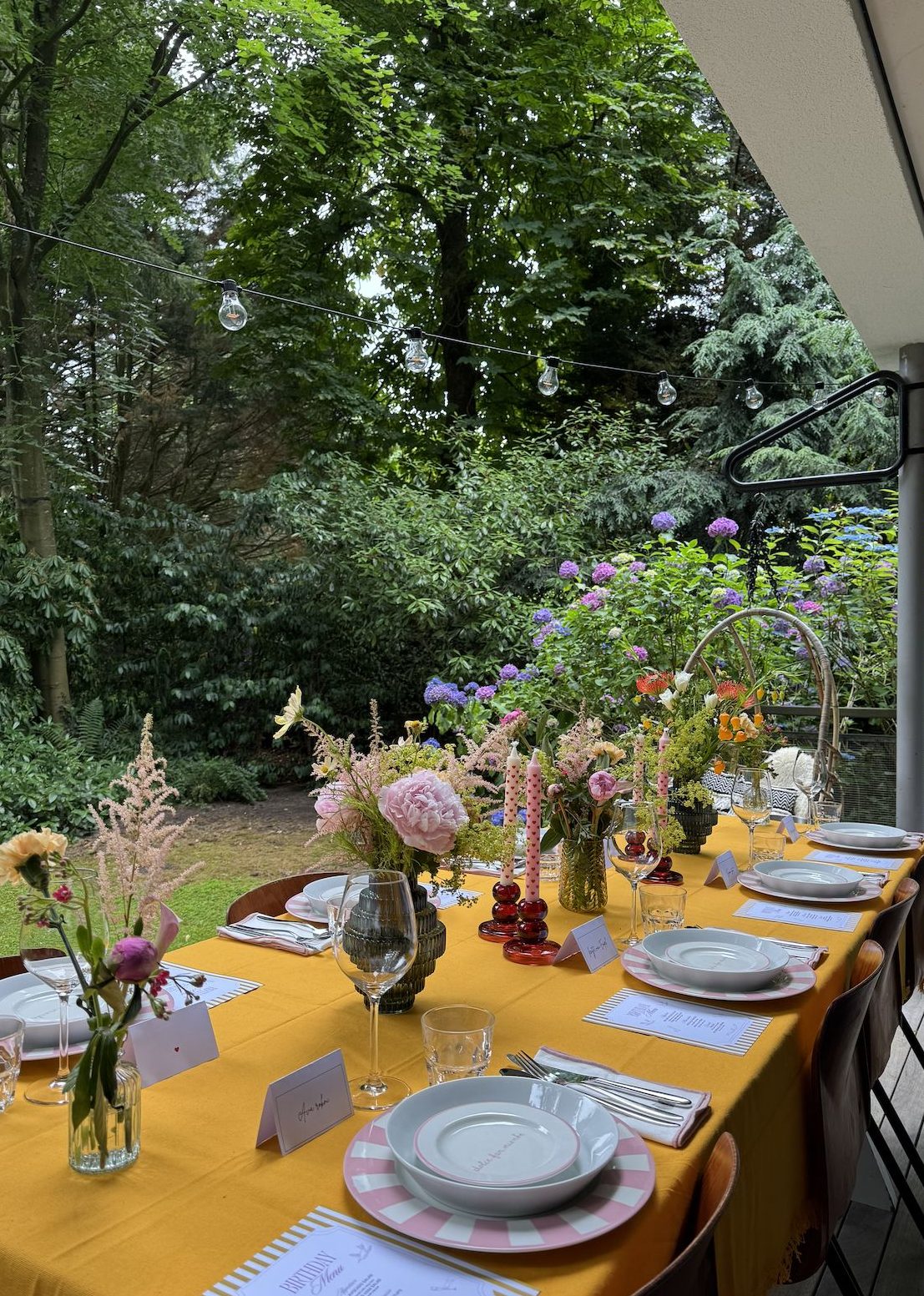 Long garden dinner table dressed in mustard linen with wildflowers and candlelight, framed by string lights and blooming hydrangeas